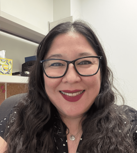A woman with long, wavy hair and glasses smiles warmly in an office setting.