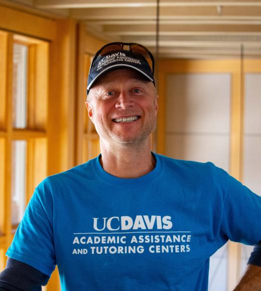 Duff Harrold in a baseball cap smiling with face and torso in frame standing inside Dutton Hall with window in the background