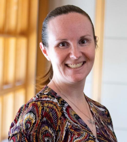 Jennifer Healey smiling with face and torso in frame standing inside Dutton Hall with window in the background