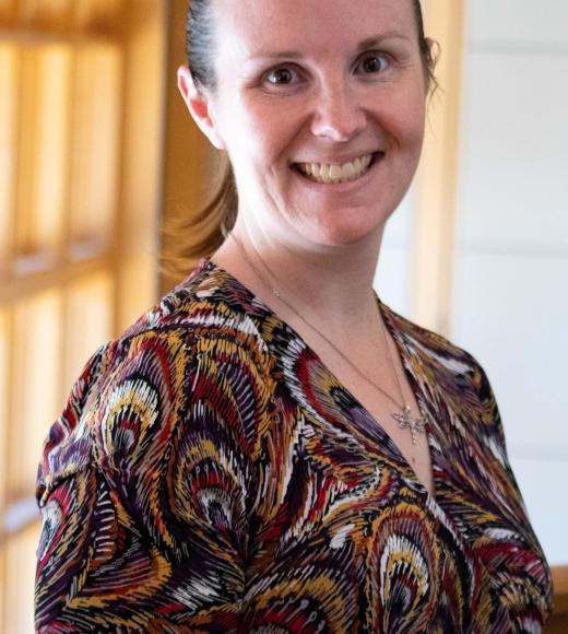 Jennifer Healey smiling with face and torso in frame standing inside Dutton Hall with window in the background